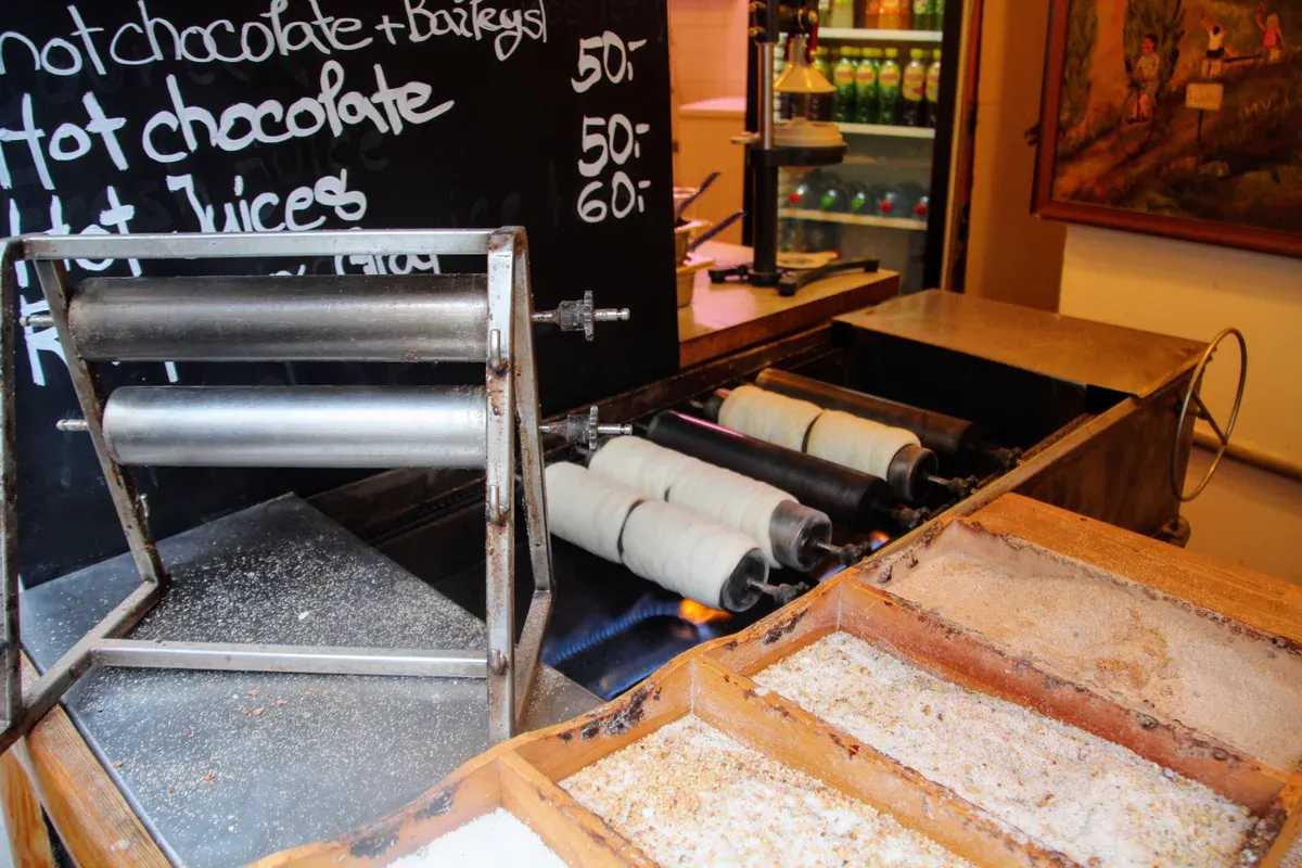 Trdelník rolls on a spit beside trays of sugar and nuts in a Prague stall
